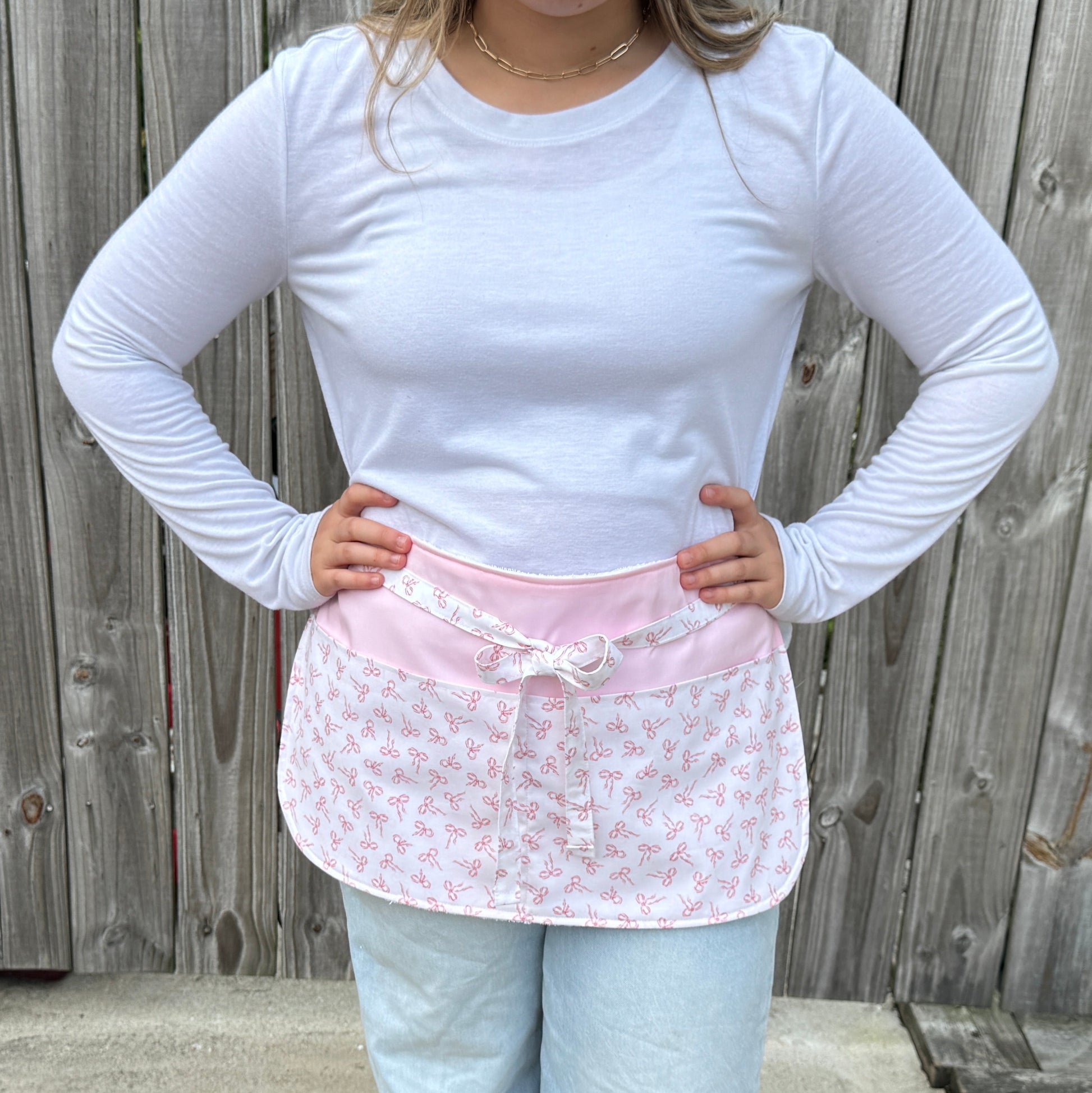 Pink and white half apron with bow print, handmade for kids, shown flat with sewing tools in pocket, cooking utensils in pocket, pdf sewing pattern for apron, easy gift to sew