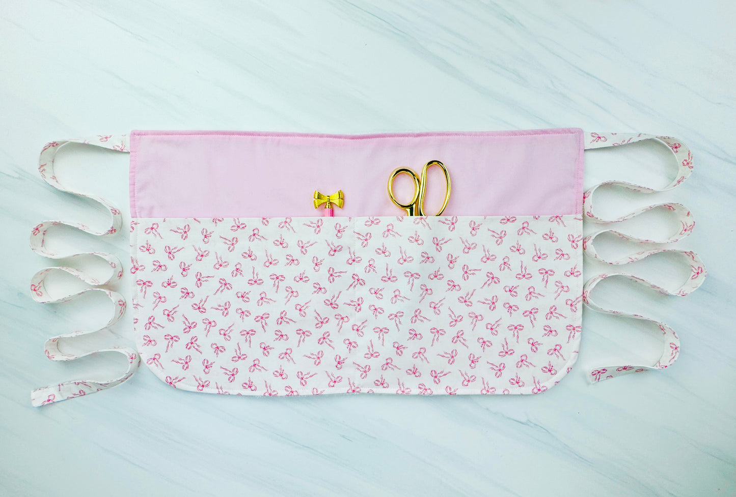 Pink and white half apron with bow print, handmade for kids, shown flat with sewing tools in pocket, cooking utensils in pocket, pdf sewing pattern for apron