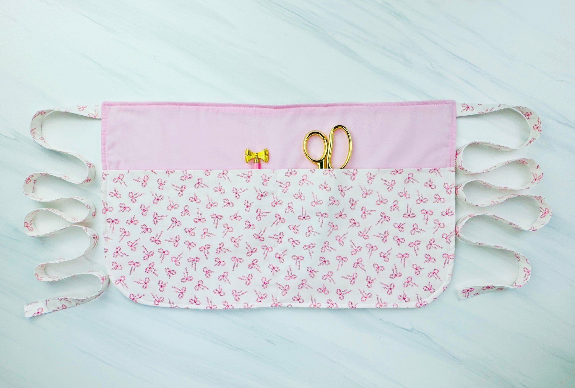 Pink and white half apron with bow print, handmade for kids, shown flat with sewing tools in pocket, cooking utensils in pocket, pdf sewing pattern for apron