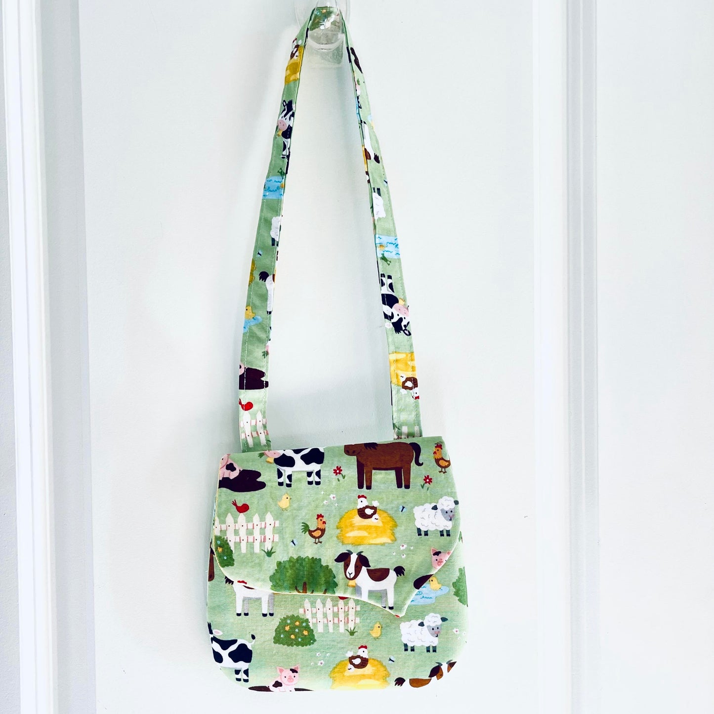 A small fabric shoulder bag with a light green background and a colorful cartoon farm animal print, hanging from a clear plastic hook on a white paneled door.