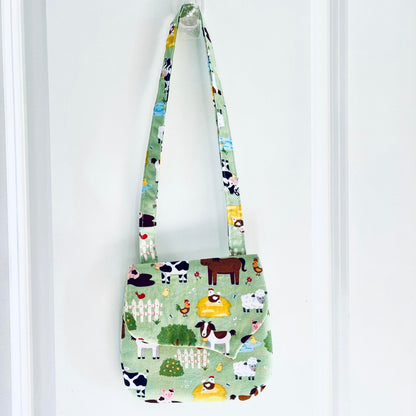 A small fabric shoulder bag with a light green background and a colorful cartoon farm animal print, hanging from a clear plastic hook on a white paneled door.