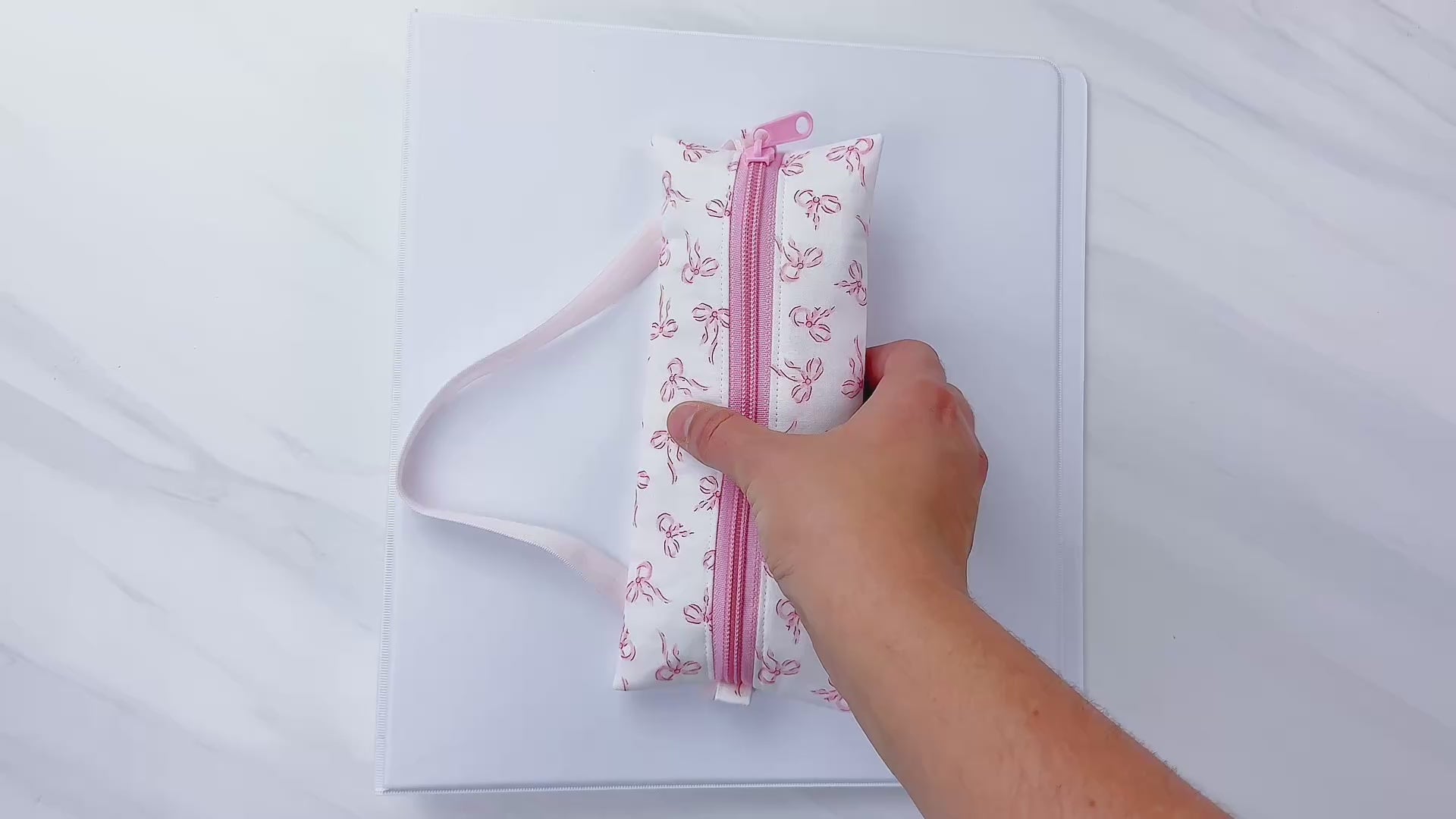 Beginner-friendly sewing pattern for a pink zippered pencil pouch with elastic, shown wrapped around school notebooks and a binder—perfect back-to-school supply case for pens, markers, and glue stick, aloha sewing company, bookmark, sewing tutorial video